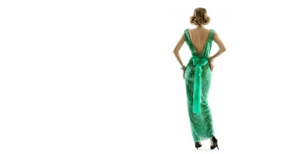 A woman in a sparkly green, backless gown with a bow stands facing away against a white background, reminiscent of a vintage dress.