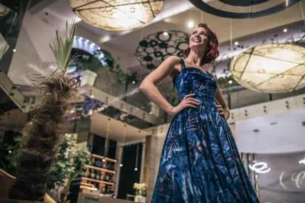 A woman in a blue cotton dress stands indoors with her hands on her hips, looking upward and smiling beneath large, circular ceiling lights.